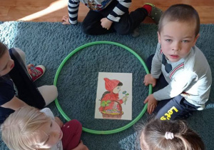 Dzieci siedzą na dywanie, odgadują: „Co nie pasuje na tym obrazku?” – na obrazku Czerwony Kapturek niesie wielkanocny koszyk.